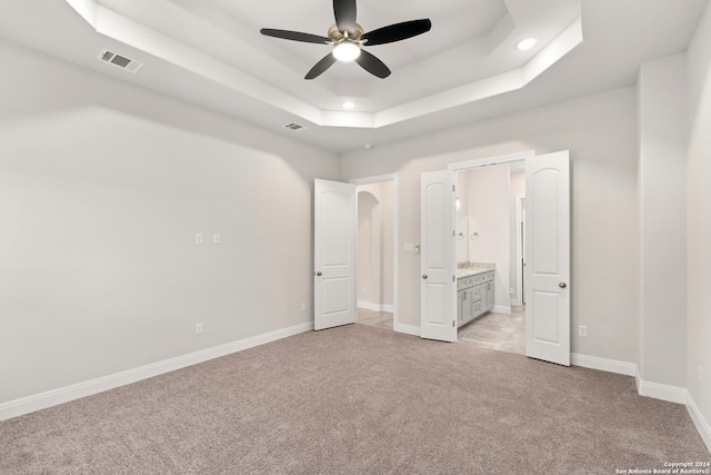 unfurnished bedroom with ceiling fan, a raised ceiling, ensuite bathroom, and light colored carpet