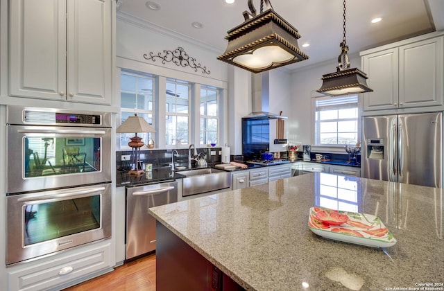 kitchen featuring light hardwood / wood-style floors, wall chimney range hood, ornamental molding, appliances with stainless steel finishes, and light stone counters