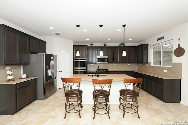 kitchen featuring hanging light fixtures, stainless steel appliances, backsplash, dark brown cabinets, and a kitchen island with sink