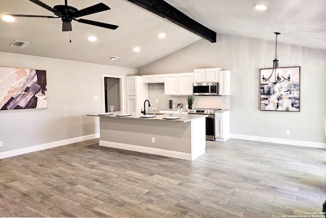 kitchen with appliances with stainless steel finishes, an island with sink, ceiling fan with notable chandelier, vaulted ceiling with beams, and white cabinets