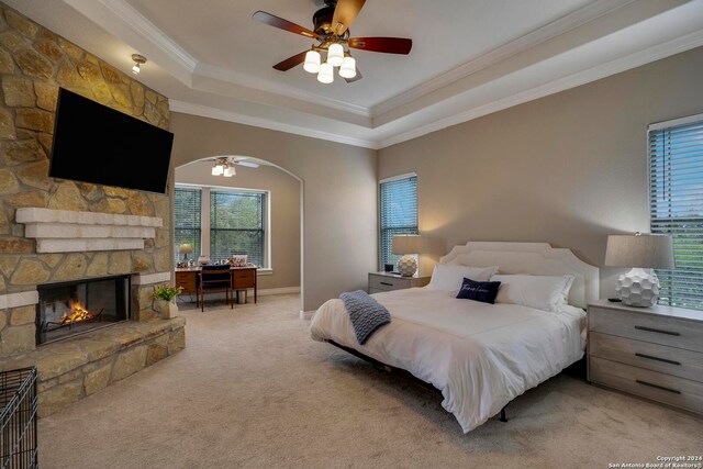 bedroom featuring ceiling fan, light carpet, a raised ceiling, a stone fireplace, and ornamental molding