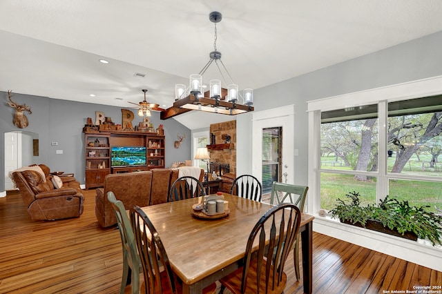 dining room with ceiling fan with notable chandelier, a stone fireplace, dark wood-type flooring, and vaulted ceiling