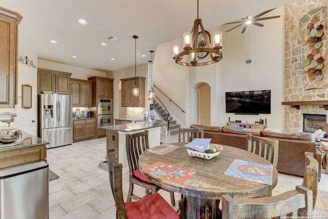 tiled dining room with sink, ceiling fan with notable chandelier, a stone fireplace, and a high ceiling