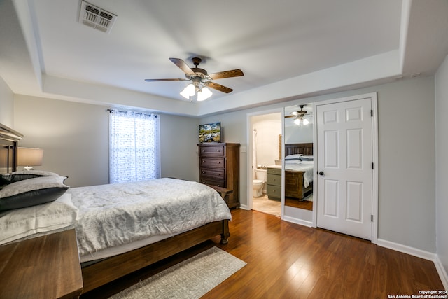 bedroom with ceiling fan, dark wood-type flooring, a tray ceiling, and ensuite bath