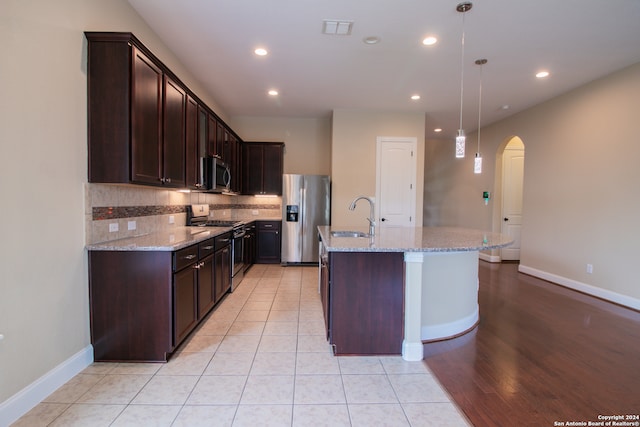 kitchen featuring an island with sink, pendant lighting, appliances with stainless steel finishes, light stone countertops, and light wood-type flooring