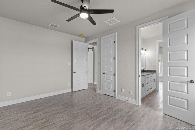 unfurnished bedroom with a barn door, light hardwood / wood-style floors, ensuite bath, and ceiling fan