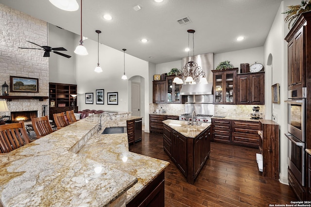 kitchen with hanging light fixtures, wall chimney exhaust hood, an island with sink, a fireplace, and sink