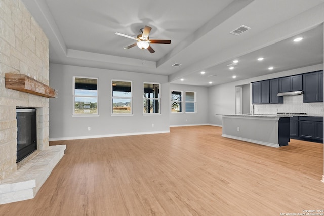 unfurnished living room featuring light hardwood / wood-style floors, a stone fireplace, ceiling fan, a tray ceiling, and sink