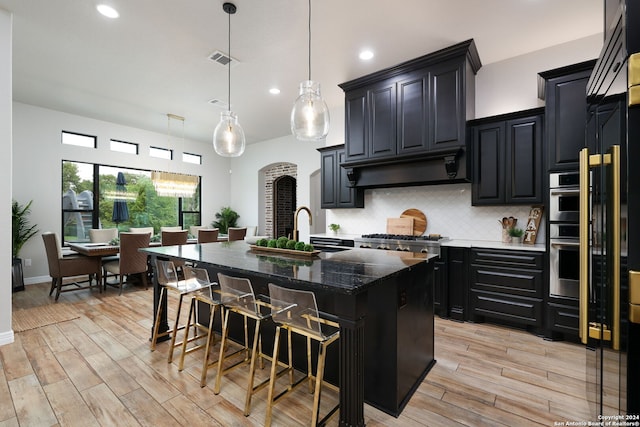 kitchen with range hood, sink, tasteful backsplash, decorative light fixtures, and a center island with sink