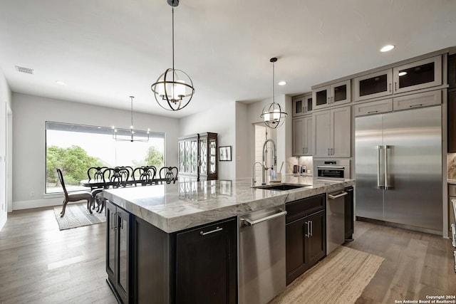 kitchen with an island with sink, stainless steel appliances, pendant lighting, light stone counters, and sink