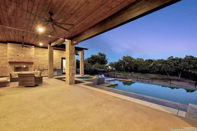 pool at dusk with ceiling fan, an outdoor living space with a fireplace, a patio, and an in ground hot tub