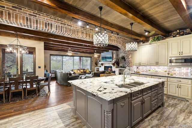 kitchen featuring a kitchen island with sink, beamed ceiling, ceiling fan with notable chandelier, stainless steel microwave, and sink