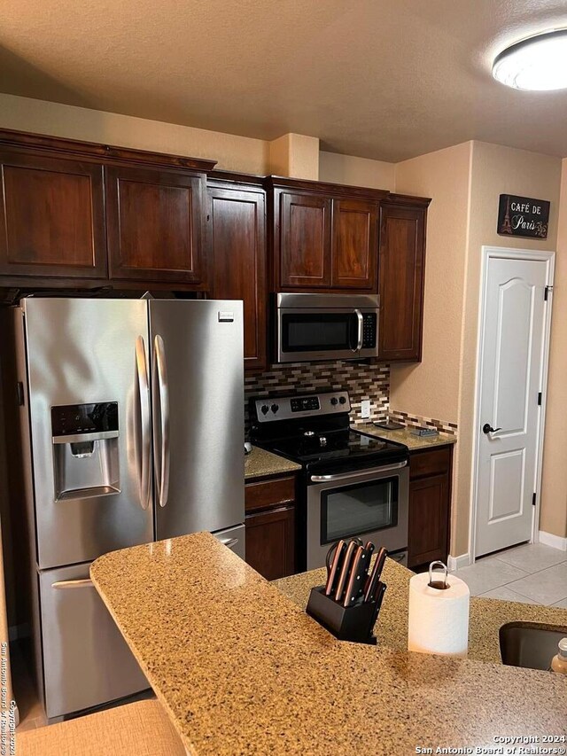 kitchen with dark brown cabinetry, light tile flooring, backsplash, stainless steel appliances, and light stone countertops