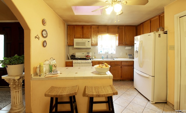 kitchen with ceiling fan, light tile floors, white appliances, tile counters, and a kitchen breakfast bar