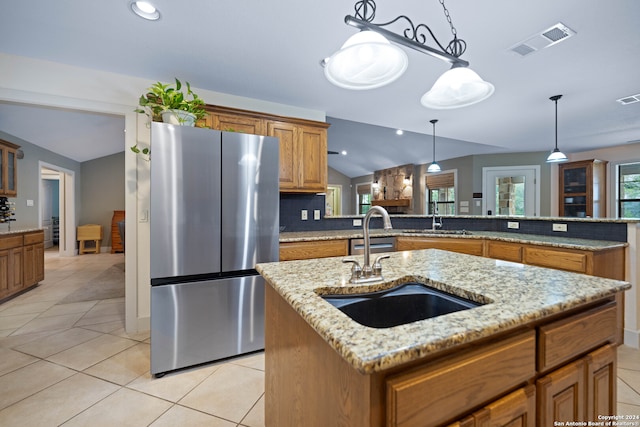 kitchen with hanging light fixtures, an island with sink, stainless steel refrigerator, vaulted ceiling, and light stone countertops