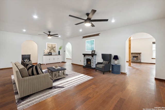 living room with a stone fireplace, dark wood-type flooring, and ceiling fan