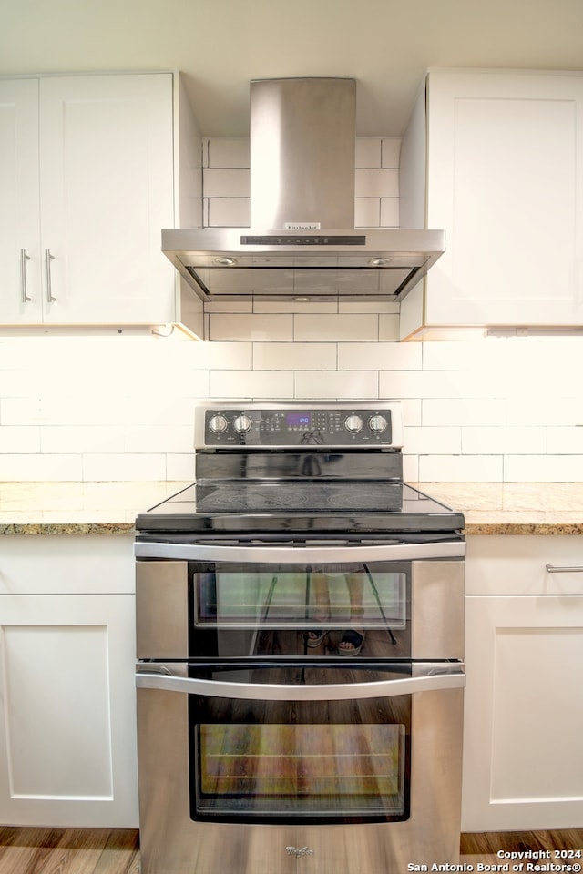 kitchen featuring wall chimney exhaust hood, tasteful backsplash, white cabinetry, range with two ovens, and light stone countertops