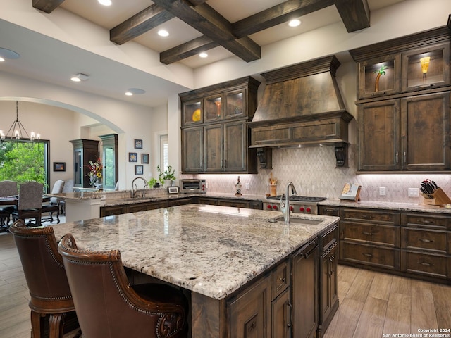 kitchen featuring pendant lighting, premium range hood, beamed ceiling, light wood-type flooring, and a center island with sink