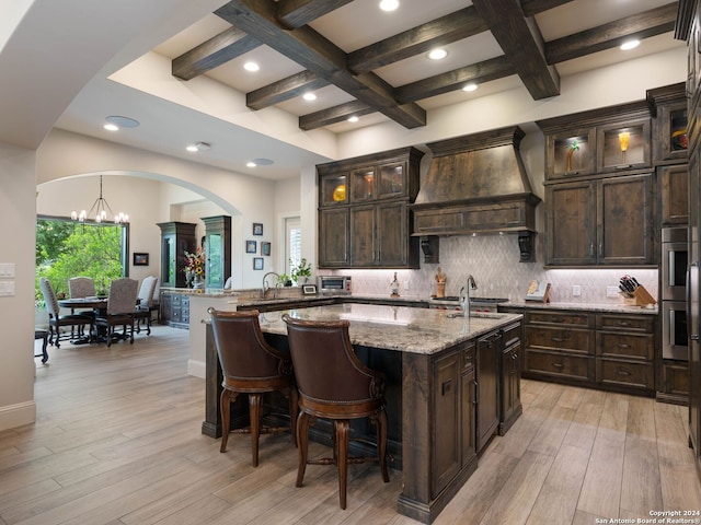 kitchen with dark brown cabinets, light stone countertops, coffered ceiling, custom exhaust hood, and light wood-type flooring