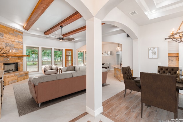 living room with ceiling fan with notable chandelier, beam ceiling, light wood-type flooring, and a stone fireplace