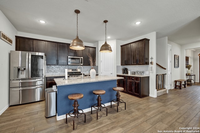 kitchen with light wood-type flooring, appliances with stainless steel finishes, an island with sink, decorative light fixtures, and tasteful backsplash