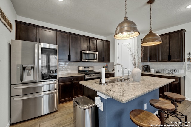 kitchen with light hardwood / wood-style floors, appliances with stainless steel finishes, an island with sink, decorative light fixtures, and backsplash