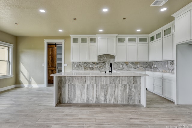 kitchen with backsplash, a barn door, a kitchen island with sink, and white cabinetry