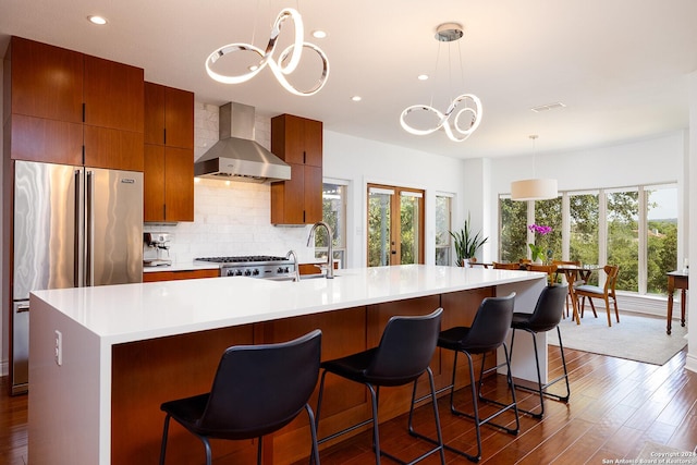 kitchen featuring high end fridge, wall chimney exhaust hood, a spacious island, sink, and hanging light fixtures