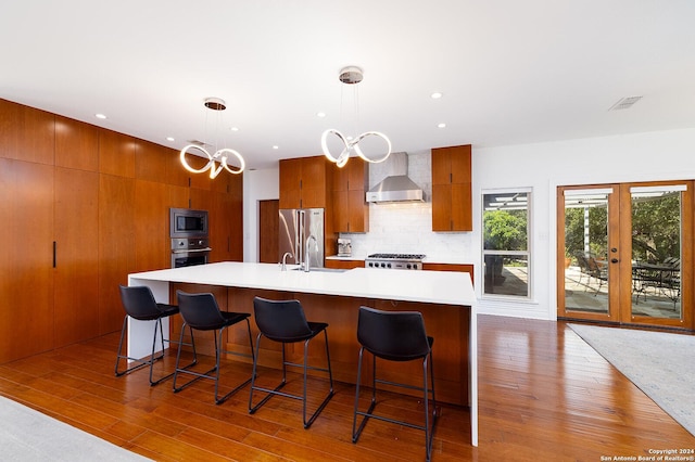 kitchen with wall chimney exhaust hood, hanging light fixtures, a large island with sink, a breakfast bar, and appliances with stainless steel finishes