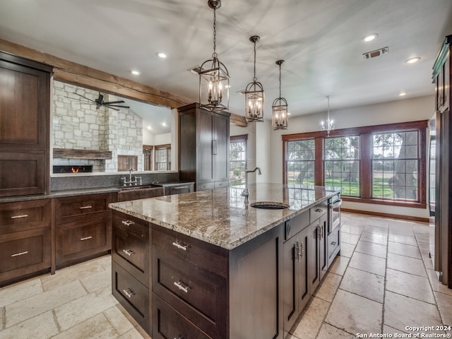 kitchen with a stone fireplace, an island with sink, light stone counters, light tile floors, and hanging light fixtures