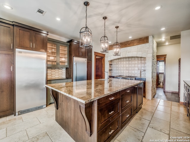 kitchen featuring stainless steel refrigerator, light hardwood / wood-style floors, hanging light fixtures, a kitchen island with sink, and built in refrigerator