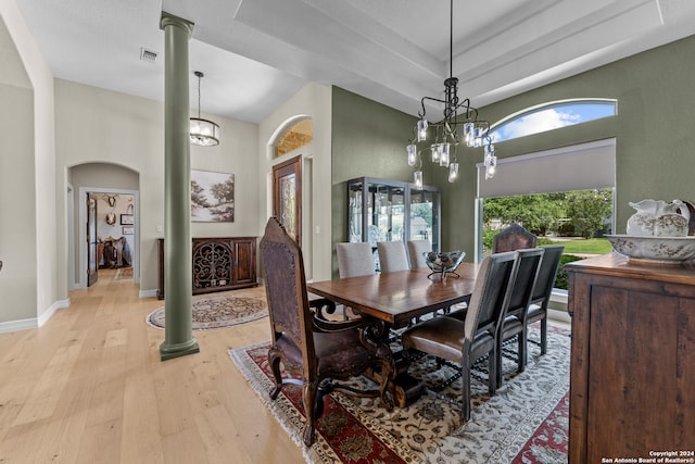 dining area featuring a towering ceiling, a notable chandelier, a tray ceiling, light wood-type flooring, and ornate columns