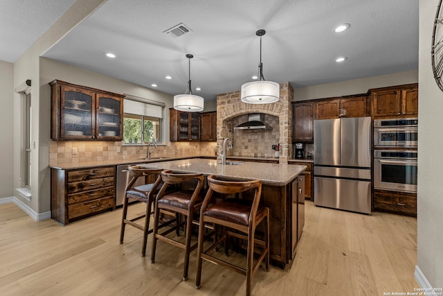 kitchen with wall chimney range hood, light hardwood / wood-style floors, stainless steel appliances, tasteful backsplash, and an island with sink