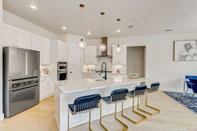 kitchen with wall chimney range hood, light wood-type flooring, a center island with sink, stainless steel appliances, and backsplash