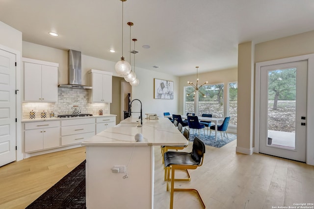 kitchen with wall chimney range hood, light hardwood / wood-style floors, an island with sink, white cabinets, and sink