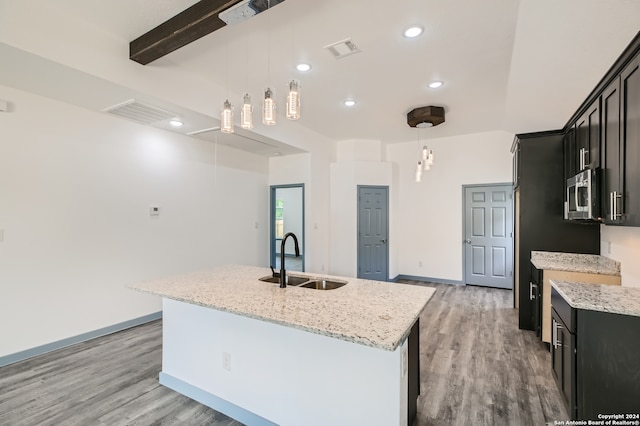 kitchen with stainless steel microwave, an island with sink, light wood-type flooring, beamed ceiling, and sink