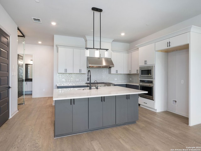 kitchen featuring tasteful backsplash, an island with sink, stainless steel appliances, light wood-type flooring, and decorative light fixtures