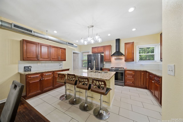 kitchen with hanging light fixtures, an island with sink, light tile flooring, wall chimney range hood, and appliances with stainless steel finishes