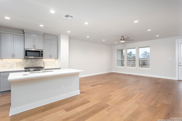 kitchen with sink, light hardwood / wood-style flooring, gray cabinetry, an island with sink, and decorative backsplash