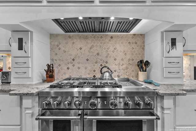 kitchen with light stone counters, backsplash, exhaust hood, white cabinetry, and double oven range