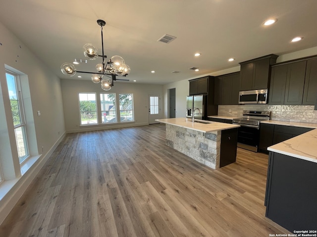 kitchen with hanging light fixtures, light wood-type flooring, light stone countertops, a kitchen island with sink, and stainless steel appliances