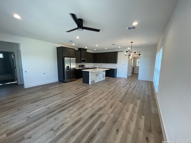 kitchen with appliances with stainless steel finishes, an island with sink, decorative backsplash, hanging light fixtures, and light hardwood / wood-style flooring