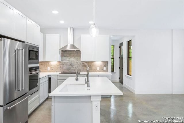 kitchen with a center island with sink, decorative light fixtures, backsplash, wall chimney range hood, and appliances with stainless steel finishes