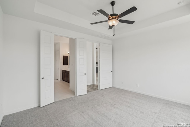 unfurnished bedroom with a raised ceiling, ensuite bath, ceiling fan, and light colored carpet