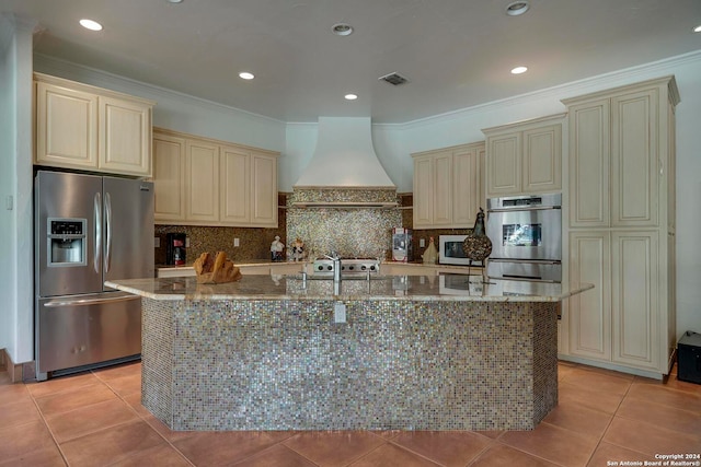 kitchen with appliances with stainless steel finishes, cream cabinets, custom range hood, and a center island with sink