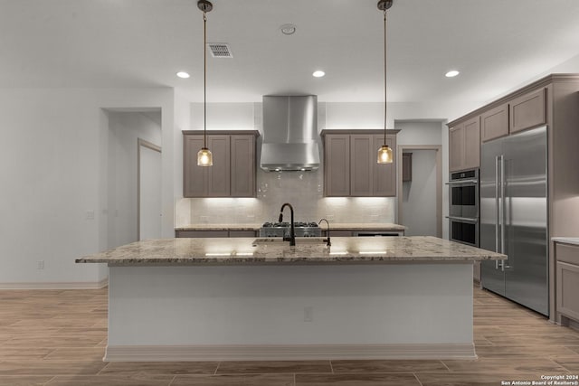 kitchen with hanging light fixtures, wall chimney range hood, an island with sink, light hardwood / wood-style floors, and appliances with stainless steel finishes