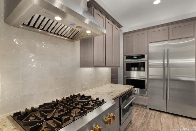 kitchen featuring backsplash, wall chimney range hood, light hardwood / wood-style flooring, light stone countertops, and stainless steel appliances