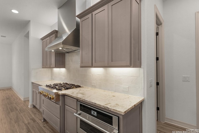 kitchen featuring wall chimney exhaust hood, light stone countertops, appliances with stainless steel finishes, tasteful backsplash, and light hardwood / wood-style floors