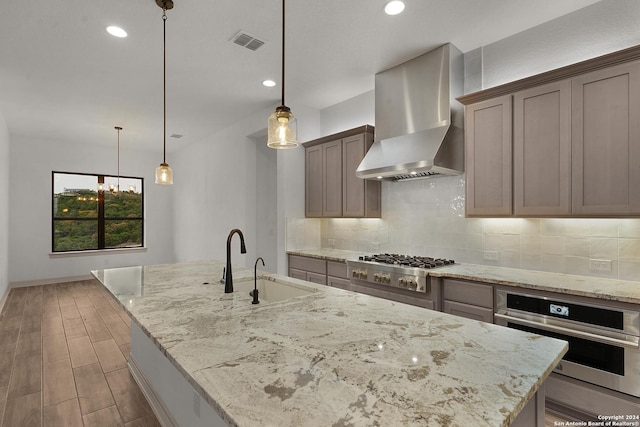 kitchen featuring light stone counters, wall chimney range hood, a kitchen island with sink, and appliances with stainless steel finishes