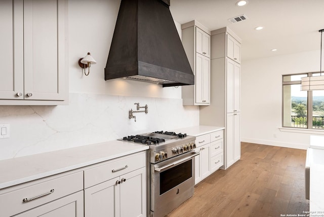 kitchen featuring custom exhaust hood, hanging light fixtures, backsplash, stainless steel stove, and light hardwood / wood-style floors